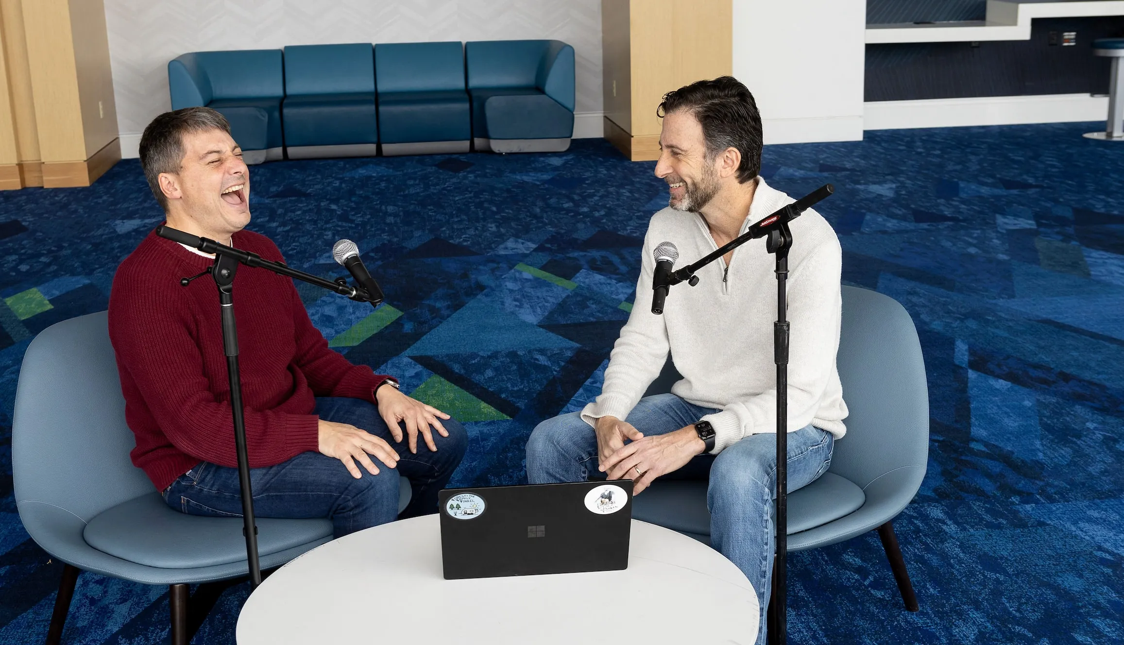 Paul Eastwick, laughing, and Eli Finkel, smiling, sit facing each other at two microphones on either side of a small round table with a laptop.